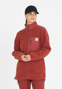 Rote Fleecejacke mit einer Reißverschlusstasche, hohem Kragen und weicher Textur; kombiniert mit roten Hosen. Verfügt über ein Logo-Patch auf der Brust.