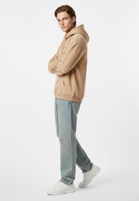 Felpa beige con cappuccio e coulisse, vestibilità rilassata. Jeans azzurri strappati e sneakers bianche. Texture morbida, design minimalista.