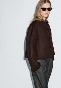 Donna che indossa occhiali da sole neri, una giacca oversize buttonata marrone scuro e pantaloni grigi a righe, in piedi contro uno sfondo chiaro e uniforme.