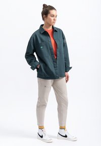 Giacca over-size verde scuro con due tasche anteriori, camicia arancione sotto, pantaloni beige e scarpe bianche con accenti neri.