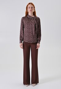 Blusa marrone con pois rosa chiaro e colletto a fiocco, abbinata a pantaloni marroni a zampa d'elefante e scarpe a punta chiare.