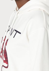 Sudadera blanca con un gráfico de tigre rojo y negro. Tiene una capucha con cordones y una tela suave y texturizada. El diseño incluye letras en negrita.