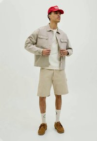 Beige denim jacket, white shirt, beige shorts, brown shoes, red cap, and white socks with green details, standing against a plain background.