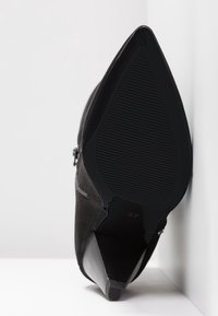 Chaussure noire à talon haut et à bout pointu avec une semelle striée, placée en angle contre une surface blanche et un mur.