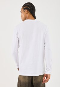 Homme avec des tresses en cornrow portant une chemise blanche à manches longues unie et un pantalon marron décoloré, de dos sur un fond blanc.