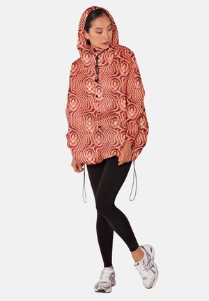 Vrouw gekleed in een rode capuchonjas met patroon, zwarte legging en witte hardloopschoenen, staand met één voet over de andere gekruist.
