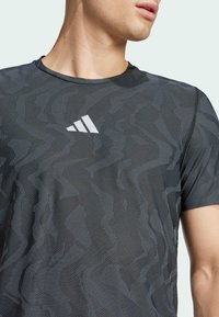 Camisa atlética negra con un patrón de zigzag y tejido texturizado. Mangas cortas, cuello redondo, con un logo blanco en el pecho.