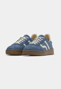 Paire de baskets en daim bleu avec lacets blancs, intérieur crème, bandes latérales blanches et semelles en caoutchouc marron sur fond gris uni.