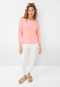 Lichtroze gebreide trui met lange mouwen en een bootnecklijn, gecombineerd met witte slim-fit broek en meerkleurige slide-sandalen.