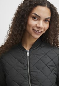 Schwarze gesteppte Bomberjacke mit Reißverschluss, geripptem Kragen und glatter Textur. Verfügt über Diamantmuster und ein silbernes Reißverschlussdetail.