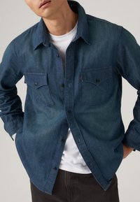 Man wearing an unbuttoned dark blue denim shirt over a white t-shirt with hands in pockets against a plain background.