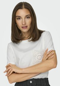 Hvid t-shirt med en tekstureret, kniplet lomme på venstre bryst, der har et rundt mønster. Modellen står med korslagte arme iført en enkel sort nederdel.