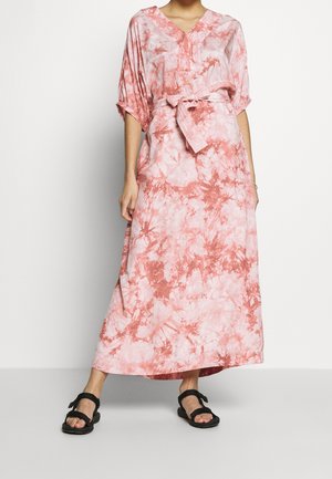 Frau trägt ein locker sitzendes Maxikleid in Pink- und Weiß-Tie-Dye mit Taillengürtel und schwarzen offenen Sandalen, steht vor weißem Hintergrund.