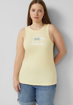 Jeune femme aux longs cheveux portant un top sans manches jaune pâle avec le texte "TUTTO BENE" et un motif de chaise de plage et parasol, associé à un short en jean.