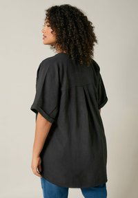 Maglietta nera oversize con maniche corte arrotolate e dettaglio plissettato sul retro. Realizzata in tessuto testurizzato, presenta un orlo arrotondato e una vestibilità ampia.
