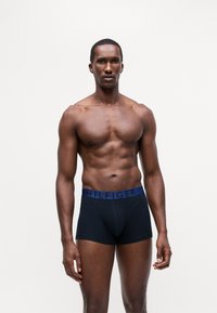 Boxeri negri cu un brâu de culoare albastră marină, având logo-ul "HILFIGER", fabricați dintr-un material neted și elastic. Modelul are o constituție musculară.