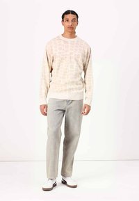 Beiger Pullover mit strukturiertem Muster, Rundhalsausschnitt und langen Ärmeln, kombiniert mit hellgrauen, taillierten Jeans und weißen Sneakers.