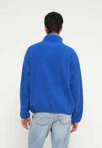 Kék polár pulóver magas gallérral, laza szabással és elasztikus szegéllyel. Világoskék farmerrel viselve, hátul a polár anyag textúrája látható.