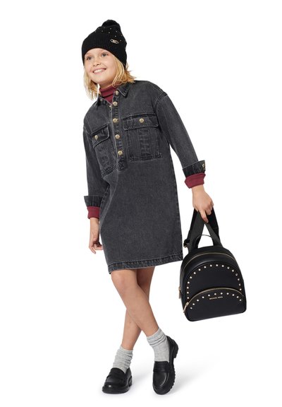 Leende barn med svart mössa, mörk denimklänning, grå strumpor och svarta skor, som håller i en svart nitarryggsäck i remmen.