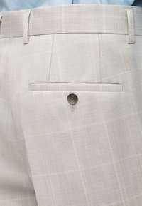 Ανοιχτό μπεζ καρό παντελόνι με πίσω τσέπη που κλείνει με ένα σκούρο κουμπί, φορεμένο με ένα ανοιχτό μπλε πουκάμισο που είναι γυρισμένο μέσα.