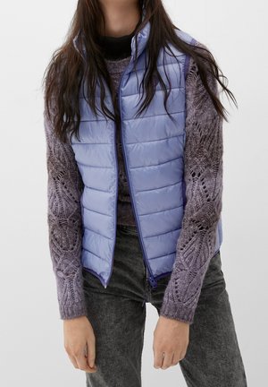 Femme portant un gilet matelassé violet clair sur un pull en tricot violet à motifs et un jean gris foncé, sur un fond uni.
