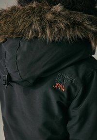 Čierna bunda s kapucňou zateplenou kožušinou. Obsahuje vyšívané "SUPER DRY JPN." v čiernej a oranžovej farbe. Materiál sa zdá byť vodeodolný.