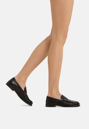 Gambe snelle che indossano mocassini in pelle nera con tacchi bassi, un piede sollevato come se camminasse, su sfondo bianco.