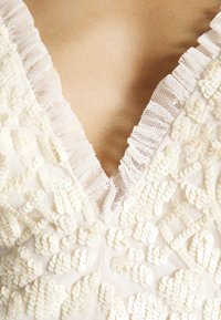 Gros plan sur une robe blanche en dentelle avec des bords froncés et des paillettes blanches, montrant un décolleté en V profond.