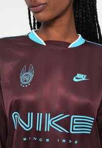 Bordo Nike majica z svetlo modrimi detajli, ki ima okrogel izrez, črtast vzorec, logo in stilizirano besedo "NIKE".