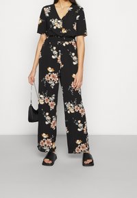 Mono negro de flores con mangas cortas, escote en V, cintura ceñida y pantalones de pierna ancha, que presenta patrones florales en rosa y blanco, llevado con sandalias negras.