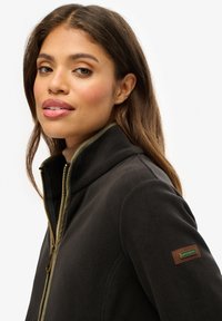 Czarna kurtka polarowa z wysokim kołnierzem, złotym zamkiem i zielonym akcentem. Posiada logo na rękawie oraz gładką teksturę.