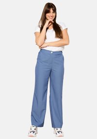 Gaiši zili plataji bikses, kas izgatavotas no teksturēta auduma, ar priekšējo pogu aizdari un sānu kabatām. Valkātas ar baltu T-kreklu un sporta apaviem.