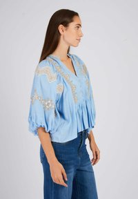 Lichtblauwe blouse met pofmouwen, een V-hals en ingewikkelde gouden borduursels op het lijfje. Uitlopende stijl met geplooide details aan de zoom.
