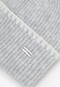 Šedá pletená čepice s žebrovanou texturou, ozdobená třásněmi a stříbrnou obdélníkovou štítkem s nápisem "WITTCHEN".