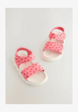 Rozā bērnu sandales ar sirdīšu rakstiem, regulējamiem Velcro siksnām un baltu teksturētu zoli labākai saķerei. Izstrādātas ērtībai un stilam.