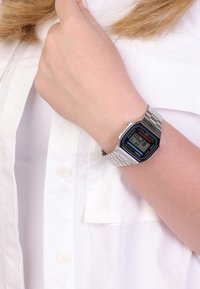 Montre en acier inoxydable argenté avec un cadran rectangulaire, accents noirs et bleus, affichage numérique indiquant l'heure, portée sur une main contre une chemise blanche.