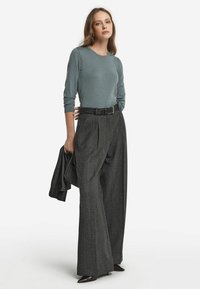 Ljusblå långärmad tröja som matchas med högmidjade, vida gråmönstrade byxor. Ett svart bälte och en matchande jacka hålls.