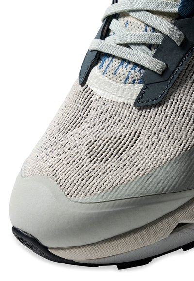 Gros plan sur la pointe d'une basket beige et gris clair avec une tige en mesh, des lacets blancs, des œillets gris foncé et une semelle texturée.