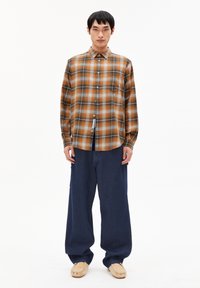 Bruine en oranje geruite overhemd met lange mouwen, gecombineerd met donkerblauwe wijde broek en beige moccasin schoenen.