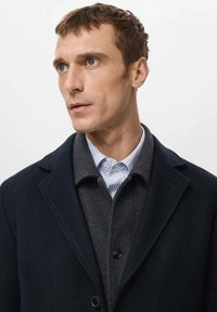 Abrigo de lana azul marino sobre un suéter gris oscuro, superpuesto a una camisa blanca con rayas azules. Se observan texturas suaves y solapas estructuradas.