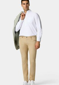 Bílá košile s dlouhými rukávy a kapsou, béžové kalhoty, světle šedý blazer přehozený přes jedno rameno a bílé tenisky. Hladká látka, přizpůsobený střih.