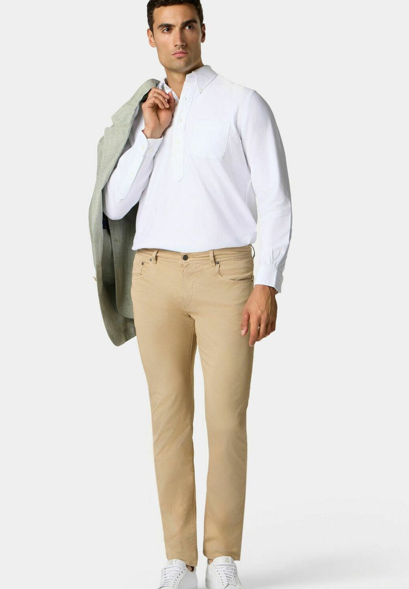 Bílá košile s dlouhými rukávy a kapsou, béžové kalhoty, světle šedý blazer přehozený přes jedno rameno a bílé tenisky. Hladká látka, přizpůsobený střih.