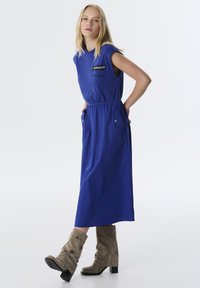 Blaues ärmelloses Kleid mit elastischem Bund, seitlichen Taschen und Logo-Detail. Tragen mit strukturierten beigen kniehohen Stiefeln und einer glatten Oberfläche.