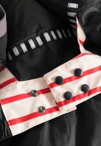 Schwarze Stoffjacke mit einem krämfarbenen Kragen, der rote Streifen aufweist. Enthält schwarze Knöpfe und ein Druckknopfverschluss-Detail.
