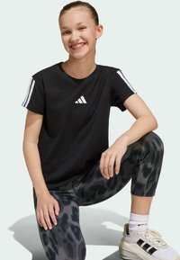 T-shirt de sport noir avec logo Adidas blanc et accents de rayures sur les épaules, associé à des leggings gris à imprimé léopard et des baskets claires.