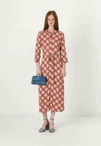 Κόκκινο φόρεμα με μοτίβο, τρία τέταρτα μανίκια, ζωνάκι στη μέση και κουμπιά. Αξεσουάρ περιλαμβάνουν μια μπλε τσάντα και συντονισμένα τυρκουάζ σανδάλια.