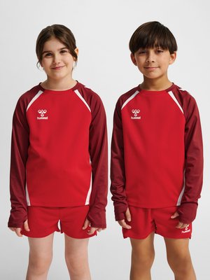 Dos niños de pie uno al lado del otro, vestidos con camisetas y pantalones cortos deportivos a juego en rojo y granate, sonriendo suavemente sobre un fondo liso.