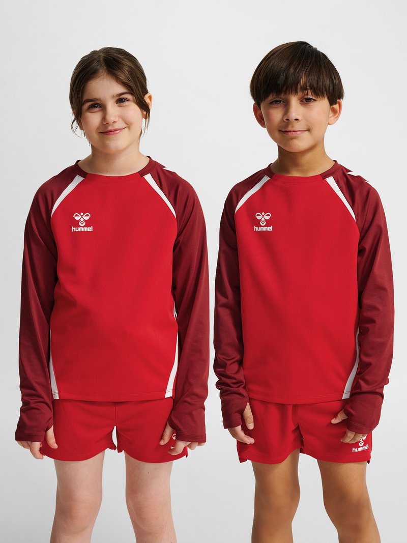 Deux enfants debout côte à côte portant des hauts et shorts de sport rouge et bordeaux assortis, souriant doucement sur un fond uni.