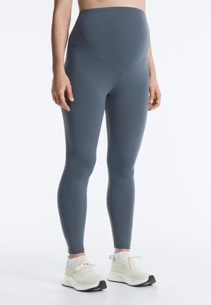 Pregnant person wearing high-waisted gray maternity leggings and white athletic shoes, standing with hands relaxed at sides.