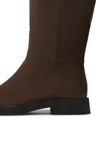 Stivale marrone in camoscio alta fino al ginocchio con suola piatta in gomma nera, texture liscia e design aderente che si estende fino al polpaccio.